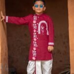 Boy standing in a traditional doorway wearing a maroon hand painted Durga motif kurta with an off-white dhoti.
