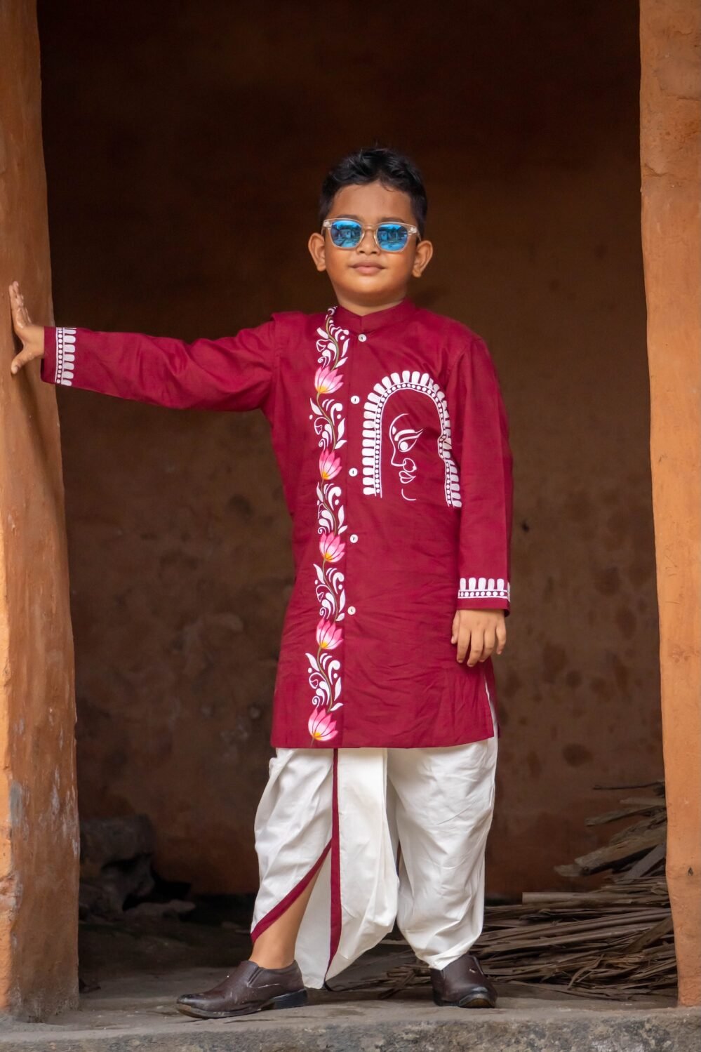 Boy standing in a traditional doorway wearing a maroon hand painted Durga motif kurta with an off-white dhoti.