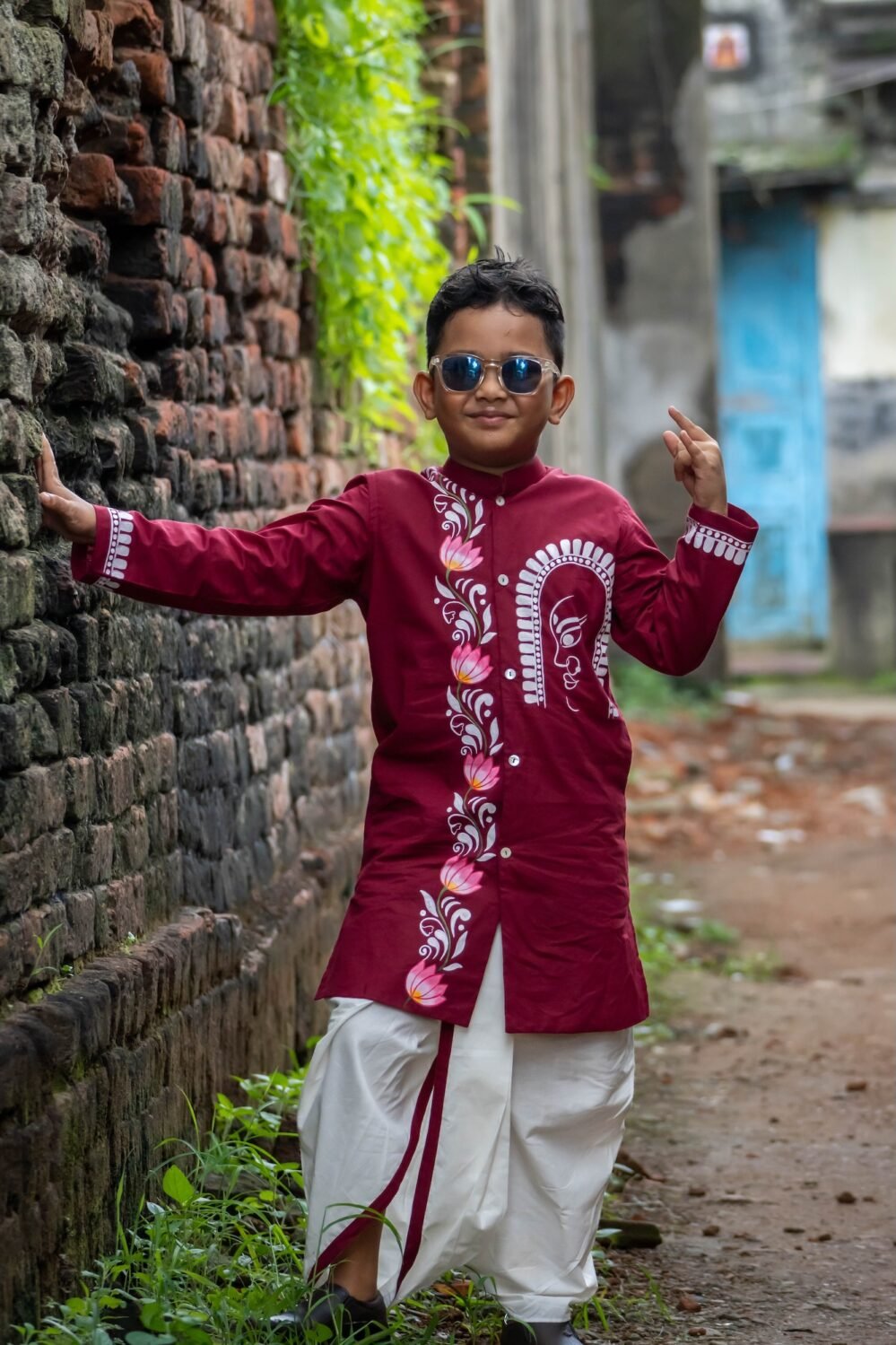 Young boy leaning against a brick wall wearing sunglasses and the Agamani maroon hand painted dhoti kurta set.