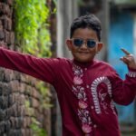 Young boy wearing a maroon hand painted Durga motif kurta by Aankibukis Couture smiling against a green metal gate backdrop.