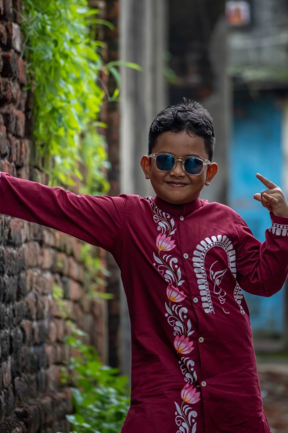 Young boy wearing a maroon hand painted Durga motif kurta by Aankibukis Couture smiling against a green metal gate backdrop.