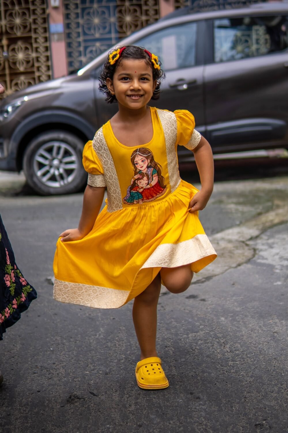 Young girl striking a playful one-legged pose wearing the yellow handpainted Durga Dulali frock with brocade border.