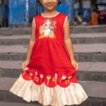 Front view of a smiling young girl holding out her red handpainted Durga Rani ethnic frock featuring a Maa Durga motif and golden frill hem.