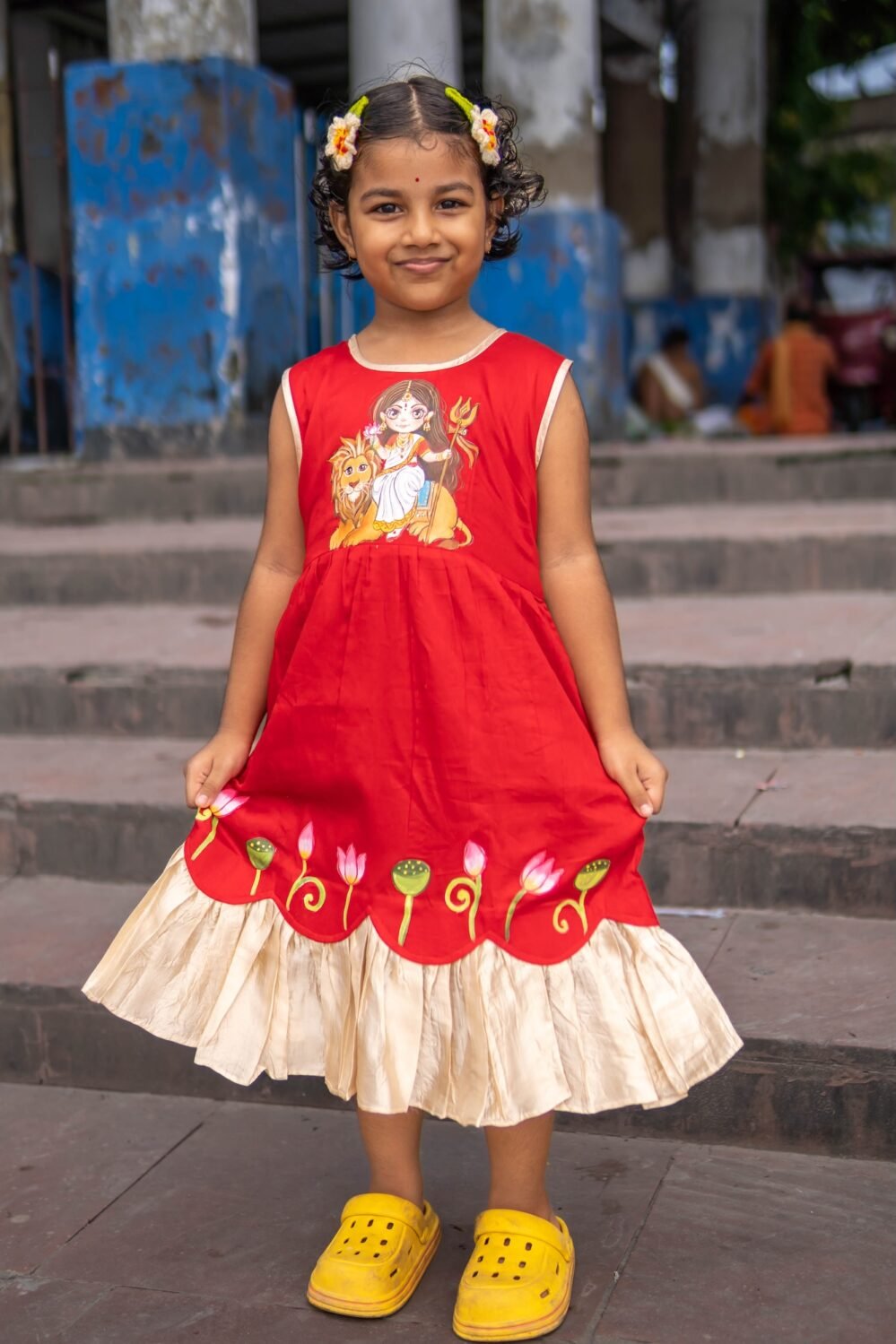 Front view of a smiling young girl holding out her red handpainted Durga Rani ethnic frock featuring a Maa Durga motif and golden frill hem.