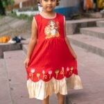 Little girl wearing red and golden handpainted Durga Rani ethnic frock holding dress skirt standing on stone steps.