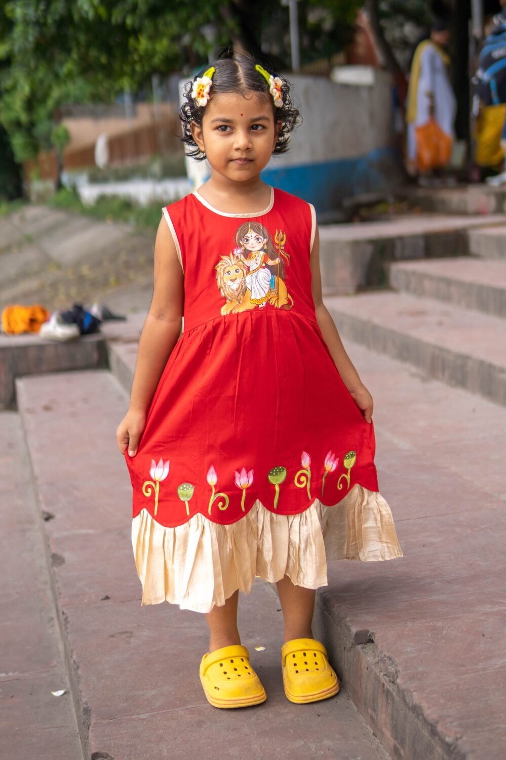Little girl wearing red and golden handpainted Durga Rani ethnic frock holding dress skirt standing on stone steps.