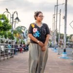 Woman posing near a park bench wearing a black blazer-collar top with hand-painted tropical flowers and beige-grey urban dhoti pants.