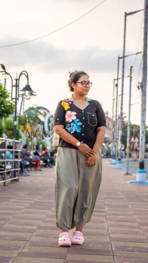 Woman posing near a park bench wearing a black blazer-collar top with hand-painted tropical flowers and beige-grey urban dhoti pants.
