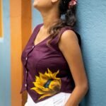 Woman wearing a purple handpainted sunflower cotton waist coat viewed from the side profile against a blue wall.