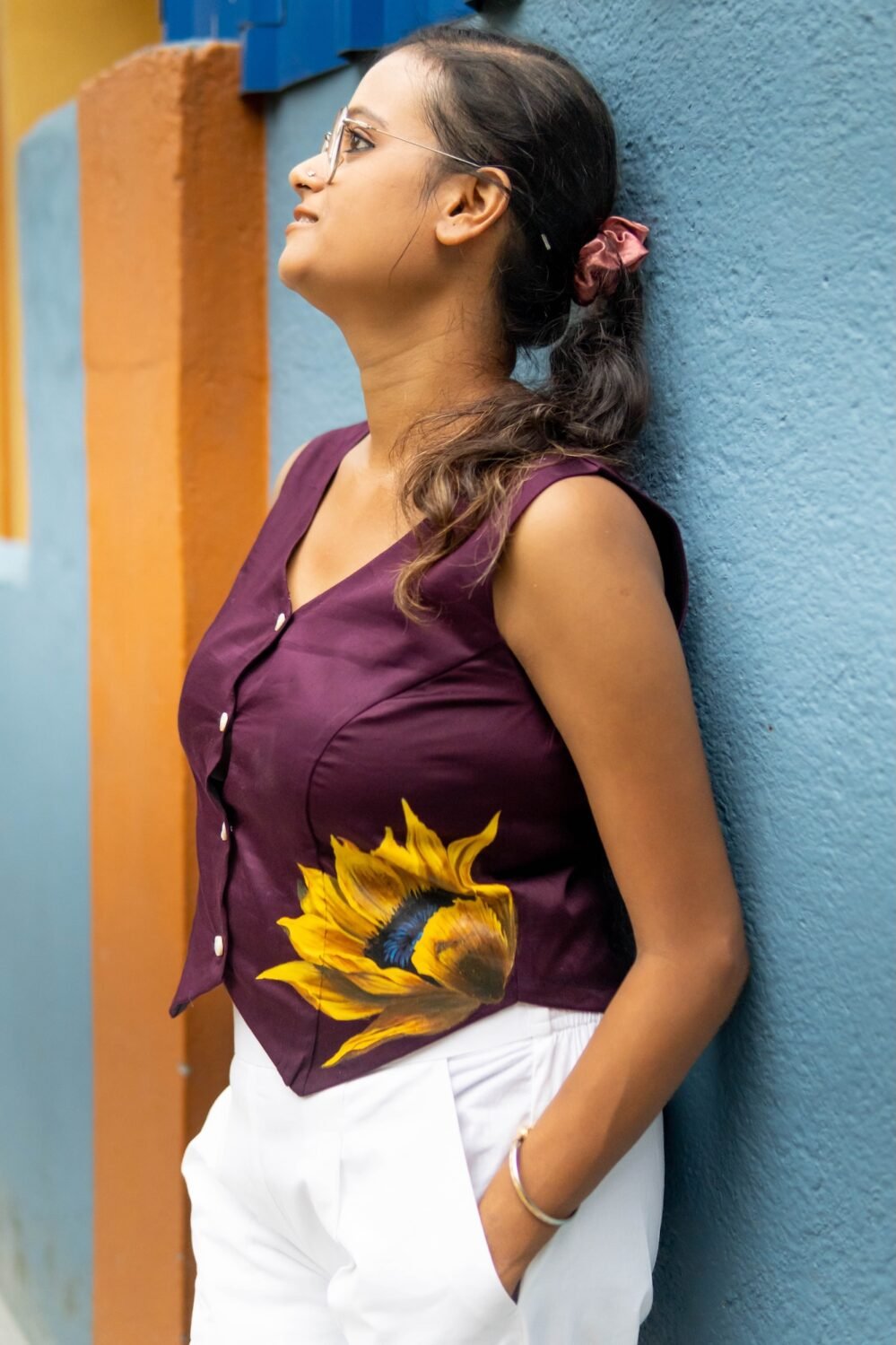 Woman wearing a purple handpainted sunflower cotton waist coat viewed from the side profile against a blue wall.