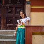 Young girl standing near temple stairs wearing hand-painted Maa Durga top and glossy peacock green festival skirt set.
