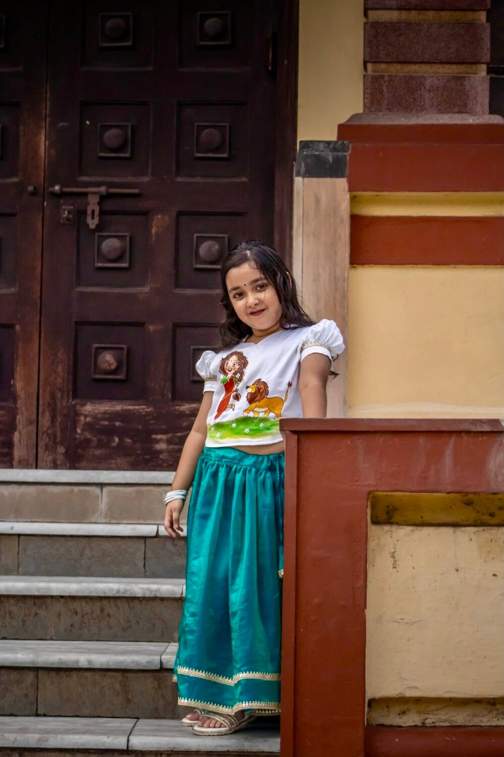 Young girl standing near temple stairs wearing hand-painted Maa Durga top and glossy peacock green festival skirt set.