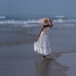 Woman walking along the ocean shoreline wearing the off-white tiered Madhubala hand-painted maxi dress with a straw hat.