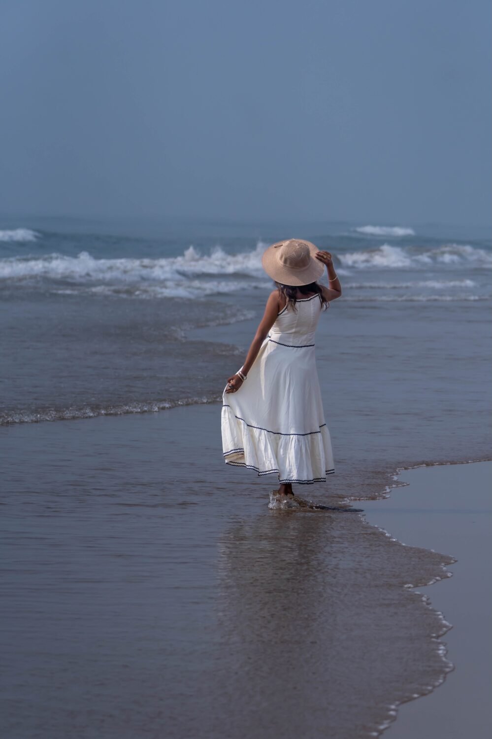 Woman walking along the ocean shoreline wearing the off-white tiered Madhubala hand-painted maxi dress with a straw hat.