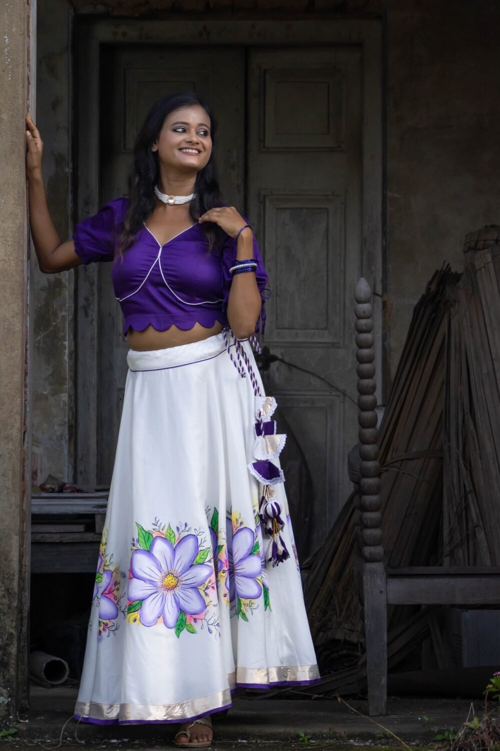 Model standing at an architectural doorway wearing the Nilaambari off-white handpainted lehenga and violet scalloped blouse set by Aankibukis Couture.