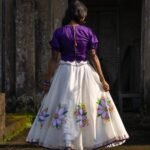 Back view of the Nilaambari deep violet puff-sleeve blouse with tie-back detail and full flared skirt with painted flowers.