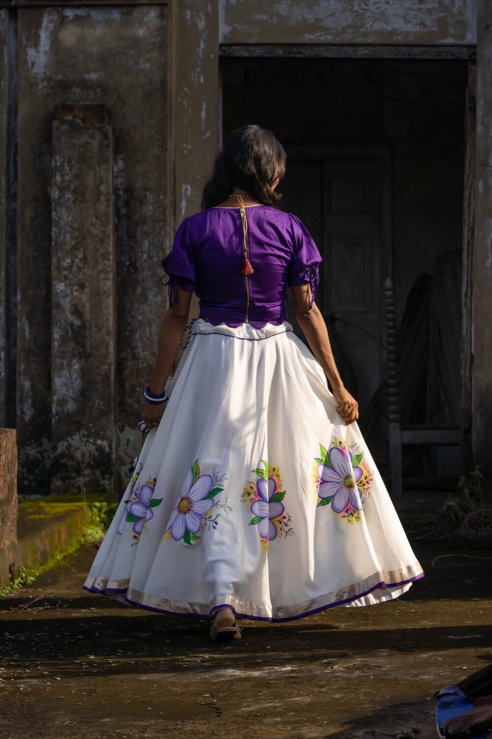 Back view of the Nilaambari deep violet puff-sleeve blouse with tie-back detail and full flared skirt with painted flowers.