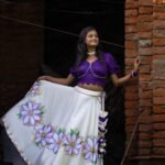 Side angle of a woman holding the flared edge of the handpainted Nilaambari off-white skirt with a rich violet crop top.