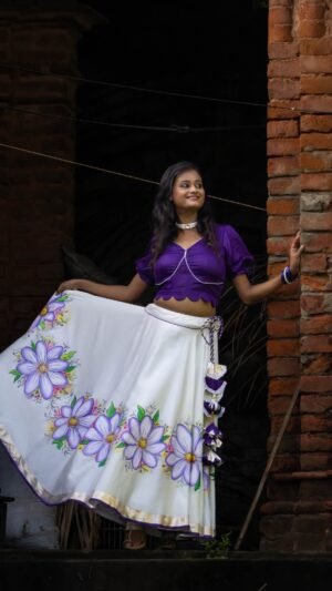 Side angle of a woman holding the flared edge of the handpainted Nilaambari off-white skirt with a rich violet crop top.