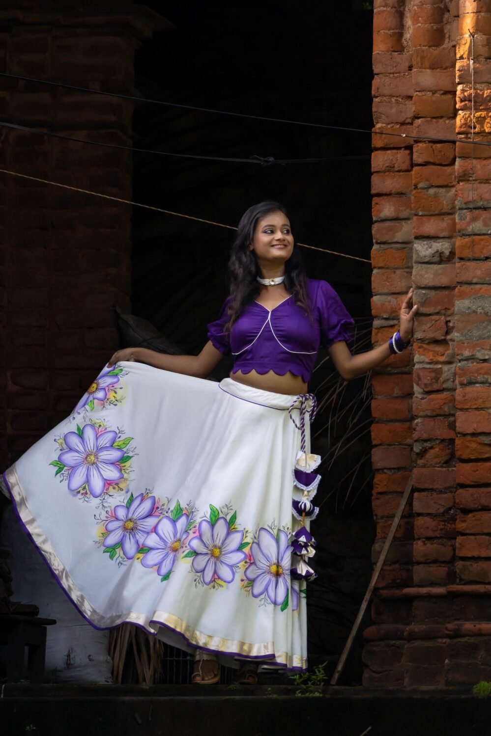 Side angle of a woman holding the flared edge of the handpainted Nilaambari off-white skirt with a rich violet crop top.