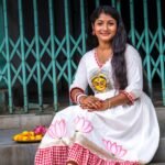 Woman sitting on marble steps wearing an off-white hand-painted Maa Durga cotton dress with pink lotus motifs and red gamcha frills.