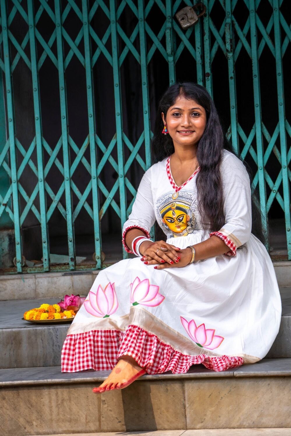 Woman sitting on marble steps wearing an off-white hand-painted Maa Durga cotton dress with pink lotus motifs and red gamcha frills.