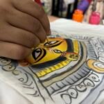 Artist hand-painting the fine eye details of the Maa Durga motif on white fabric with a thin paintbrush.