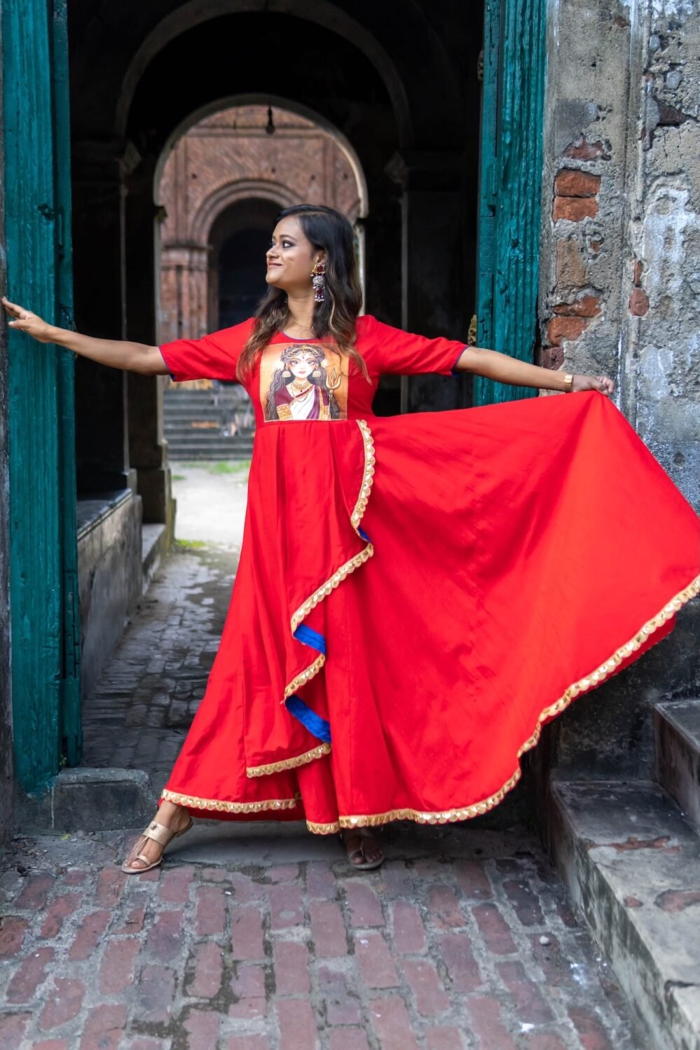 Model spreading the wide skirt flare of the hand-painted red Maa Durga gown in a doorway.