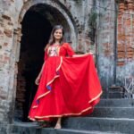 Model holding out the flare of the red Maa Durga hand-painted gown under an ancient brick archway.