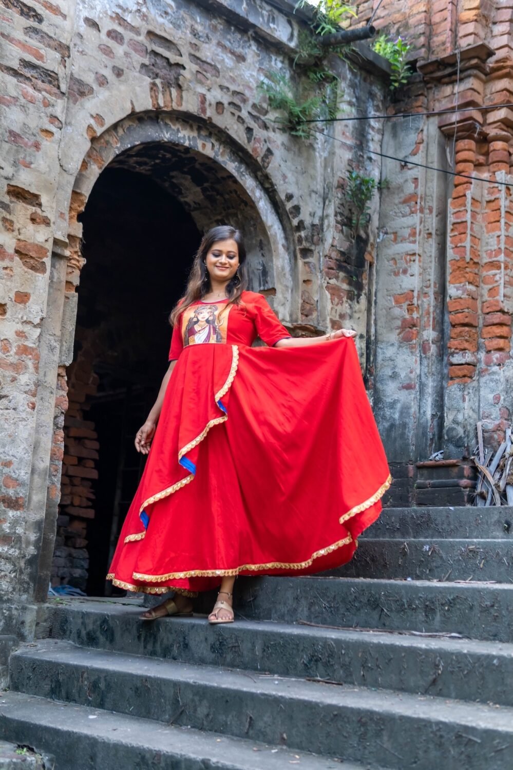 Model holding out the flare of the red Maa Durga hand-painted gown under an ancient brick archway.